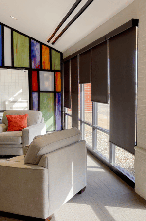 corner of bright sunny room with tan couches and dark roller shade window treatments and stained glass background